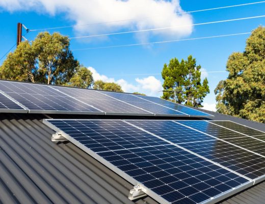 New solar panels are installed on the metal sheet roof of the house in a shaded area with tall trees on a sunny day.