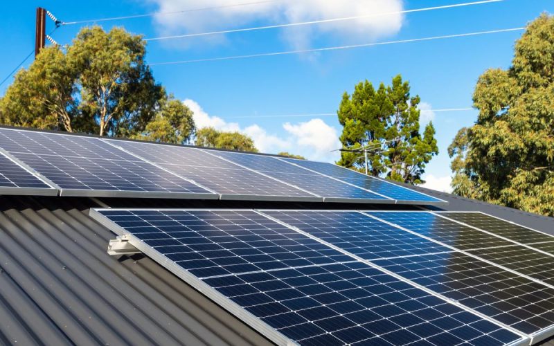New solar panels are installed on the metal sheet roof of the house in a shaded area with tall trees on a sunny day.