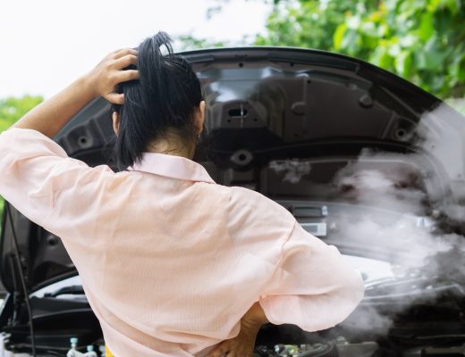 A woman standing in front of her car with the hood up. She is looking down at her smoking car engine.