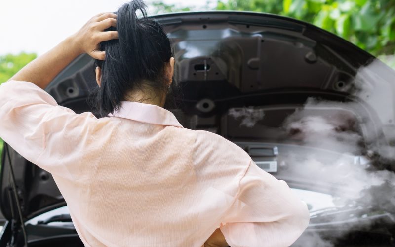 A woman standing in front of her car with the hood up. She is looking down at her smoking car engine.