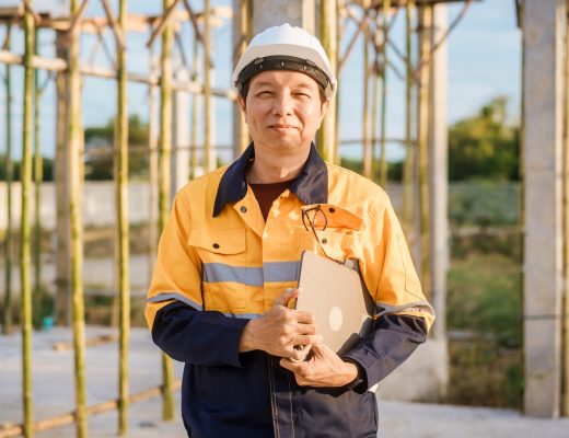 A contractor wears a white hard hat and orange safety vest while holding a tablet and standing near scaffoldings.