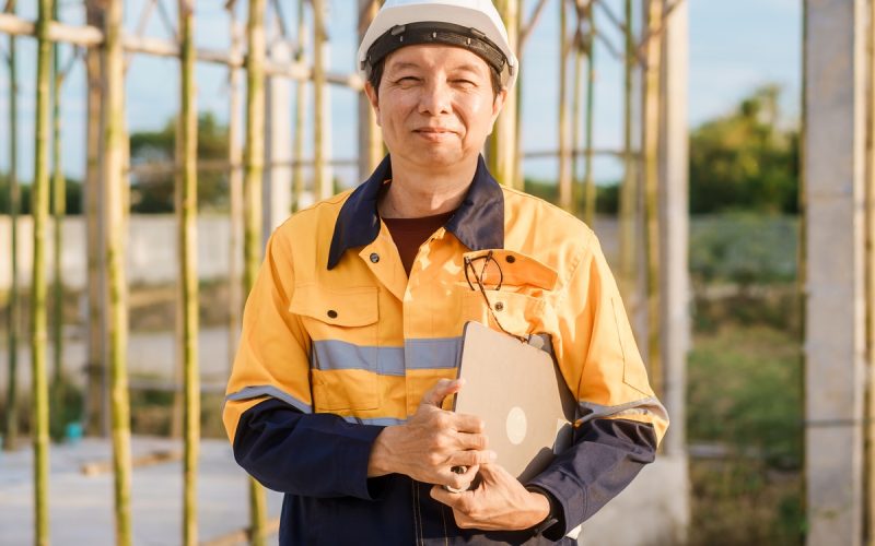 A contractor wears a white hard hat and orange safety vest while holding a tablet and standing near scaffoldings.