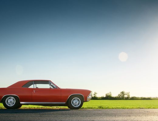 A red, classic muscle car is parked on the side of a road next to an empty field under a clear, sunny sky.