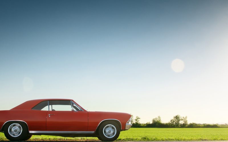 A red, classic muscle car is parked on the side of a road next to an empty field under a clear, sunny sky.