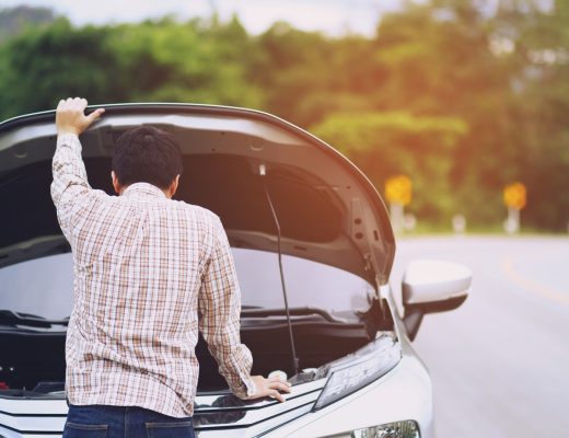 A man stands in front of his vehicle parked on the side of the road. His arm lifts the hood as he looks at the engine bay.