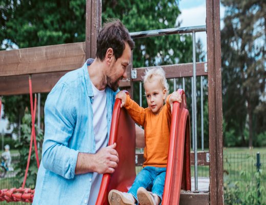 A young dad smiles as he watches his toddler prepare to slide down a red slide on an outdoor playset.