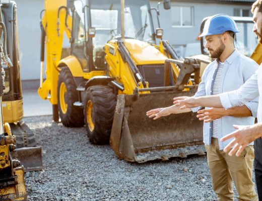 Two men looking at some construction equipment. Both are gesturing toward one of the vehicles in front of them.
