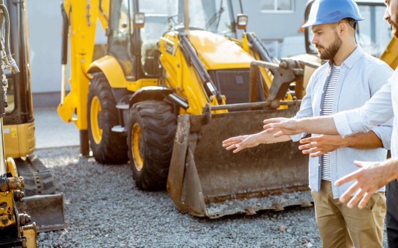 Two men looking at some construction equipment. Both are gesturing toward one of the vehicles in front of them.