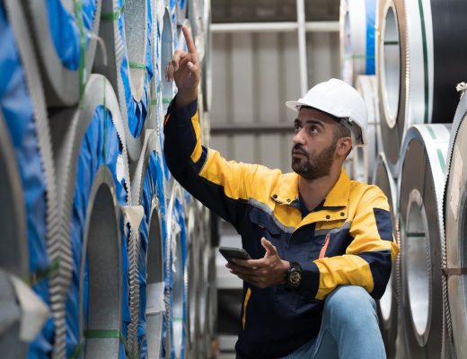 A man in a hardhat crouched next to some stacks of aluminum coils. He is looking up and pointing to one of the higher ones.