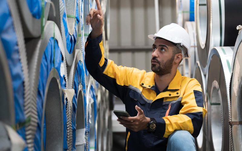 A man in a hardhat crouched next to some stacks of aluminum coils. He is looking up and pointing to one of the higher ones.