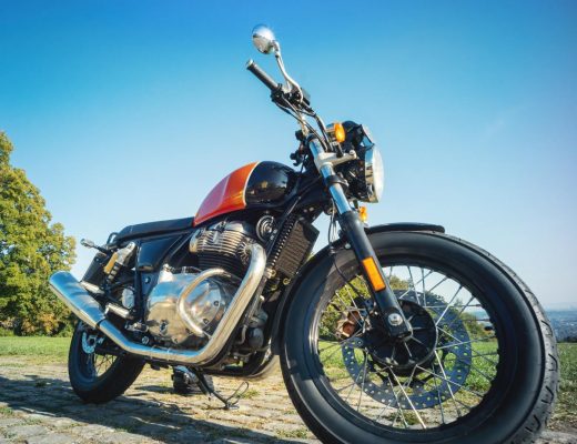 A black and orange motorcycle parked on top of a cobblestone path lined with grass. There are trees in the distance.