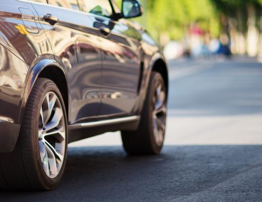 The sun reflects off a black car, casting a shadow on the road as it drives. In the distance are rows of trees.