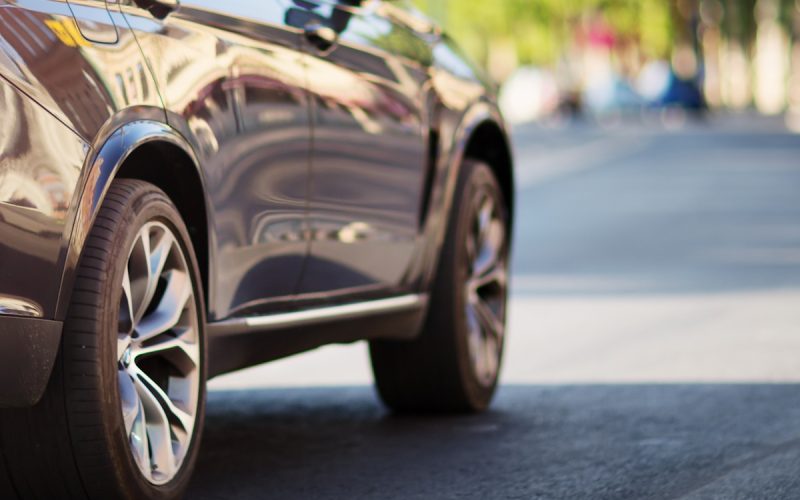The sun reflects off a black car, casting a shadow on the road as it drives. In the distance are rows of trees.