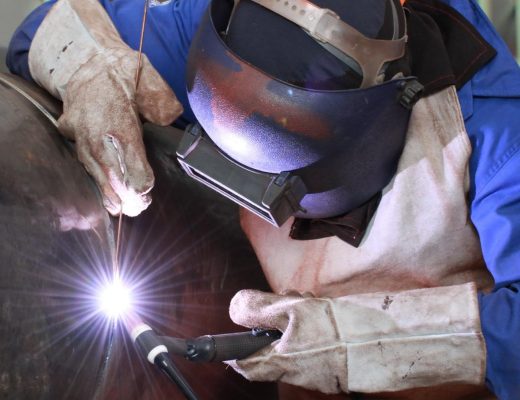 A person dressed in complete welding gear, including a helmet and gloves, using a torch on a large metal tube.