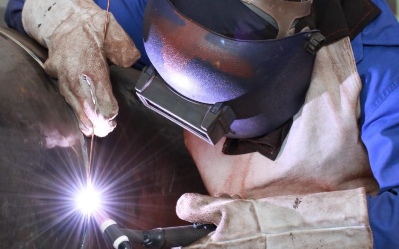 A person dressed in complete welding gear, including a helmet and gloves, using a torch on a large metal tube.