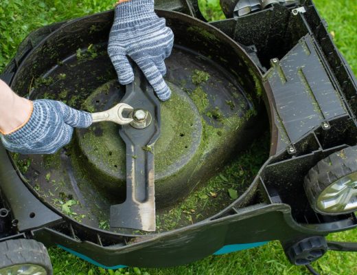 A man working on the underside of a mower. He's wearing gloves and using a wrench to tighten a bolt on the blade.
