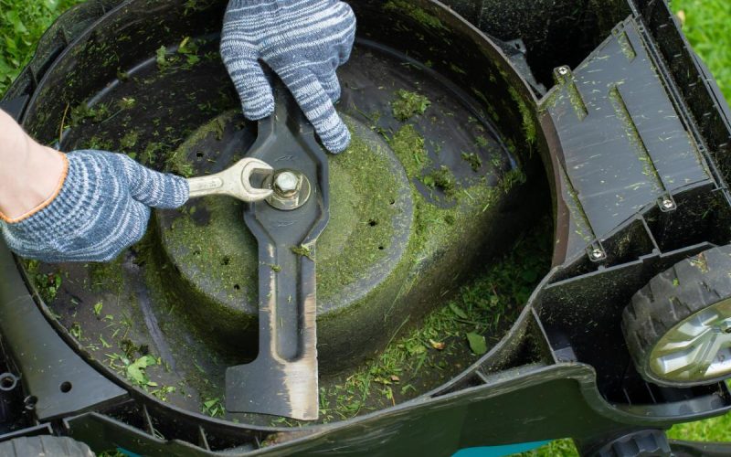 A man working on the underside of a mower. He's wearing gloves and using a wrench to tighten a bolt on the blade.
