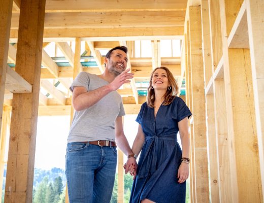 A couple is building their dream home and walking hand in hand through the wooden structure to look at the foundation.