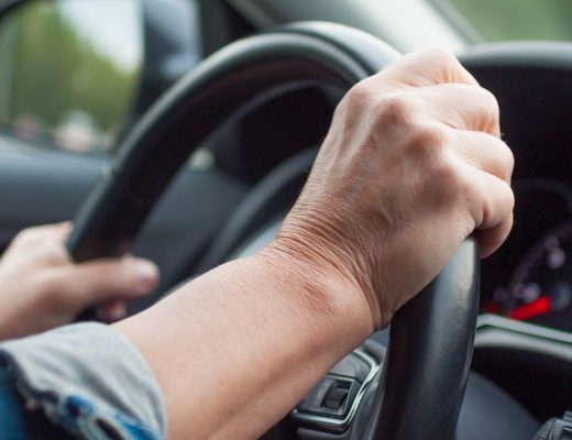 A driver grips a black steering wheel in a car, with a denim sleeve visible and a side mirror reflecting green foliage.