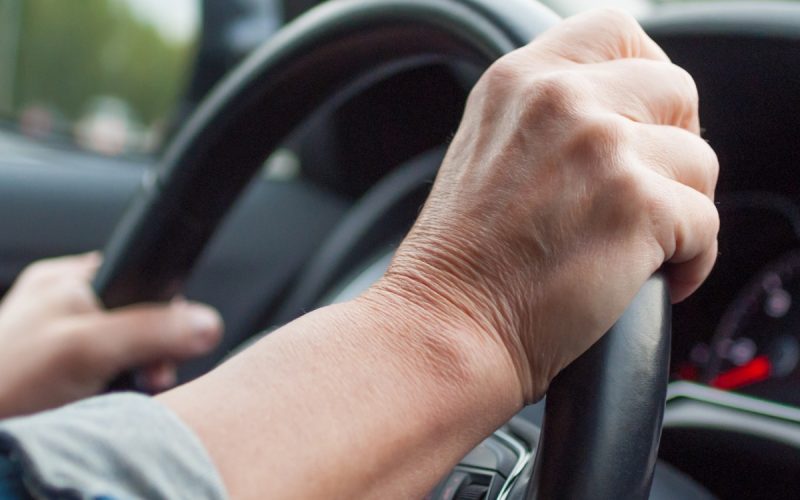 A driver grips a black steering wheel in a car, with a denim sleeve visible and a side mirror reflecting green foliage.