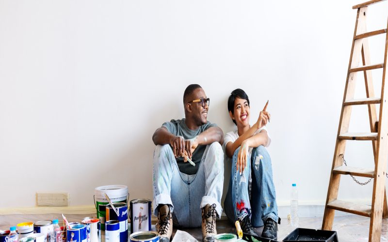 A couple is sitting down on the floor and smiling at their hard work while they take a break from painting the house wall.