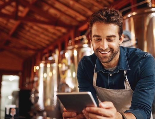 A close-up of a man, wearing an apron, smiling, while looking at a tablet in the distillery factory.