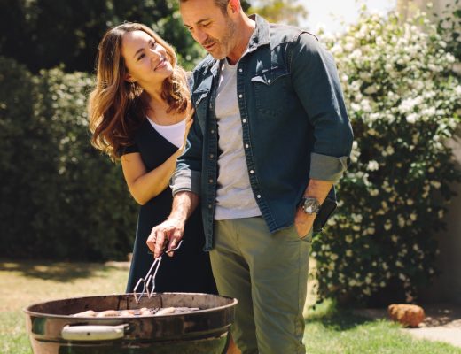 A happy couple is grilling in their backyard. The wife is hugging her husband from behind as the husband grills.