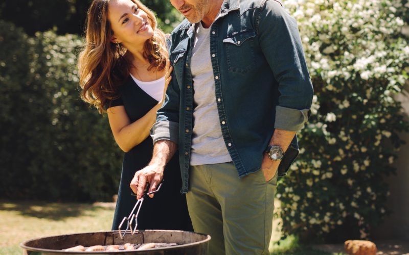 A happy couple is grilling in their backyard. The wife is hugging her husband from behind as the husband grills.