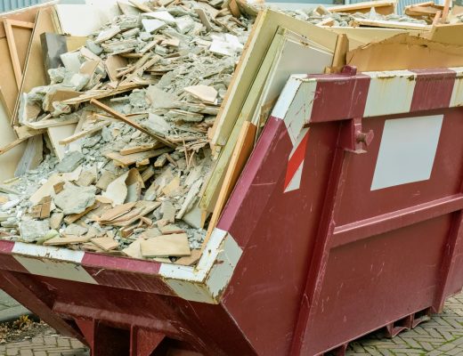 A large, red dumpster parked in a lot contains wood, drywall, and other types of debris from a construction project.