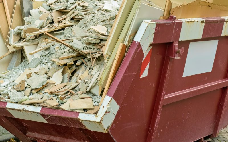 A large, red dumpster parked in a lot contains wood, drywall, and other types of debris from a construction project.