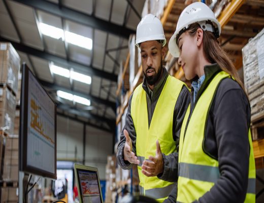 Two warehouse workers in yellow safety vests and white hard hats stand beside one another in front of a computer.