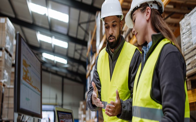 Two warehouse workers in yellow safety vests and white hard hats stand beside one another in front of a computer.