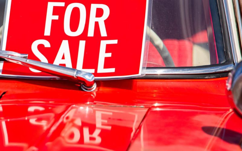 Close-up of red vehicle windshield and hood, showing a bright FOR SALE placard beside the wiper arm.