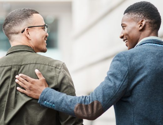 Two businessmen are walking outside a stone building. One man places his hand on the other person's back.