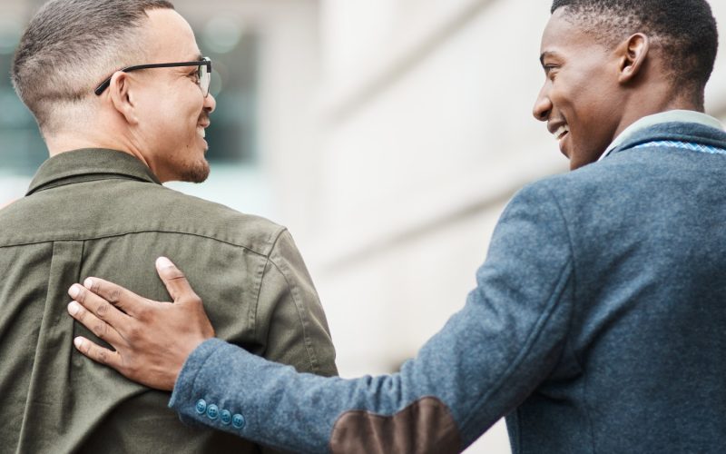 Two businessmen are walking outside a stone building. One man places his hand on the other person's back.