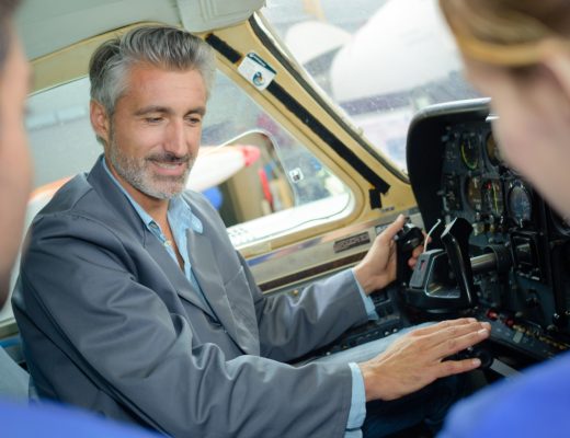 Man in airplane cockpit points to instrument panel and controls while two passengers watch from nearby seats.