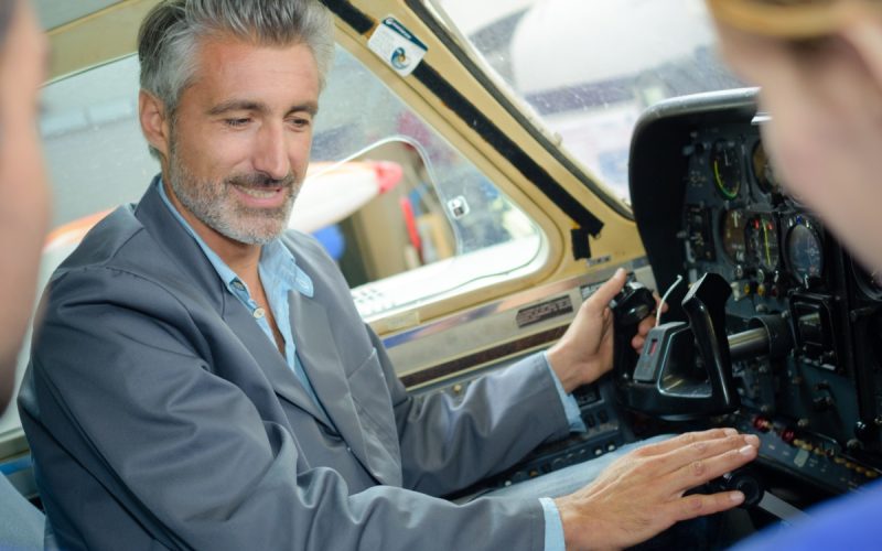 Man in airplane cockpit points to instrument panel and controls while two passengers watch from nearby seats.