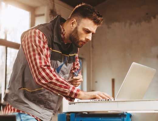 A man wearing a work jacket and holding a pair of work glasses in one hand peers at a laptop sitting on a workbench in his garage.