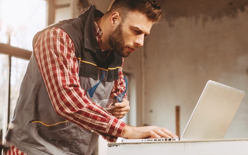 A man wearing a work jacket and holding a pair of work glasses in one hand peers at a laptop sitting on a workbench in his garage.