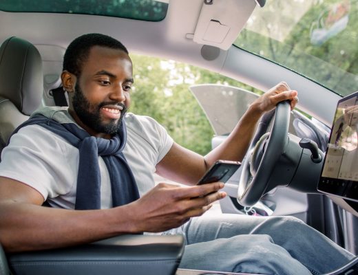 A man sits in the driver's seat of a car with the door open. He looks at his phone, with the car's infotainment display lit up.