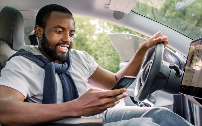 A man sits in the driver's seat of a car with the door open. He looks at his phone, with the car's infotainment display lit up.