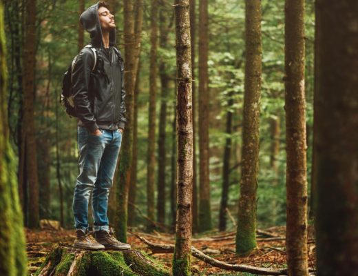 A calm man in hiking gear taking a deep breath while standing on a tree stump in the middle of a green wooded area.