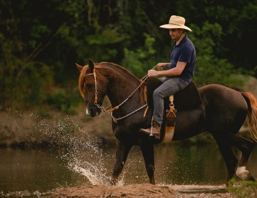 Man wearing a straw hat riding a saddled horse through a river, splashing water upward as he rides the horse.