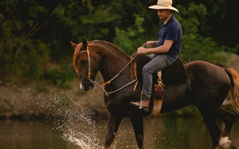 Man wearing a straw hat riding a saddled horse through a river, splashing water upward as he rides the horse.
