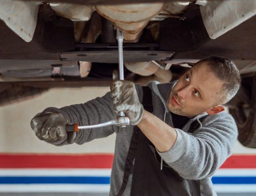 A man cranes his head to look at the undercarriage of a car on a hydraulic lift while using a torque wrench.
