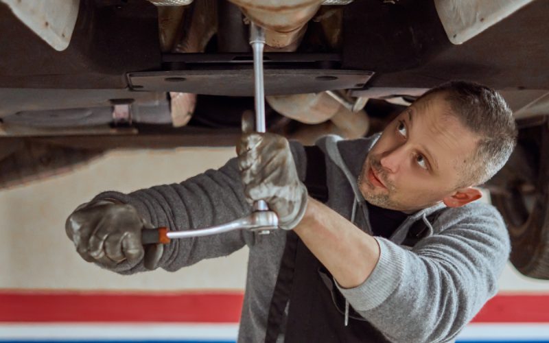 A man cranes his head to look at the undercarriage of a car on a hydraulic lift while using a torque wrench.