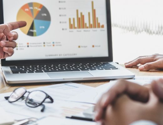 Three pairs of hands around a table full of papers. One hand is pointing at a laptop screen full of business data.