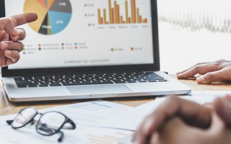 Three pairs of hands around a table full of papers. One hand is pointing at a laptop screen full of business data.