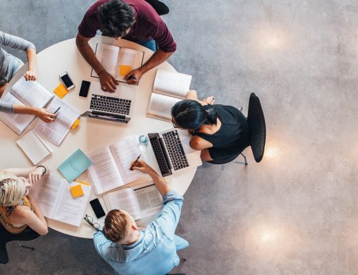 Top-down view of group studying at table with laptops, notebooks, papers, and phones arranged around workspace.