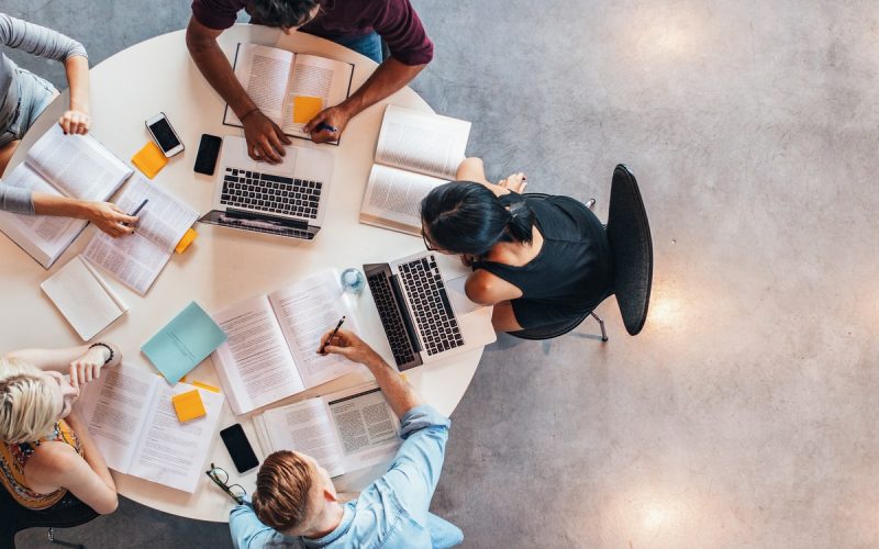 Top-down view of group studying at table with laptops, notebooks, papers, and phones arranged around workspace.